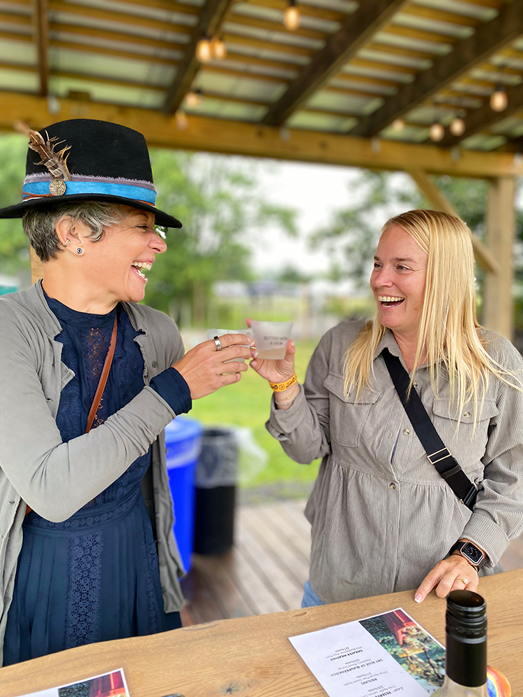 image of two women bumping cups while enjoying a wine tasting under the pergola at the concert venue